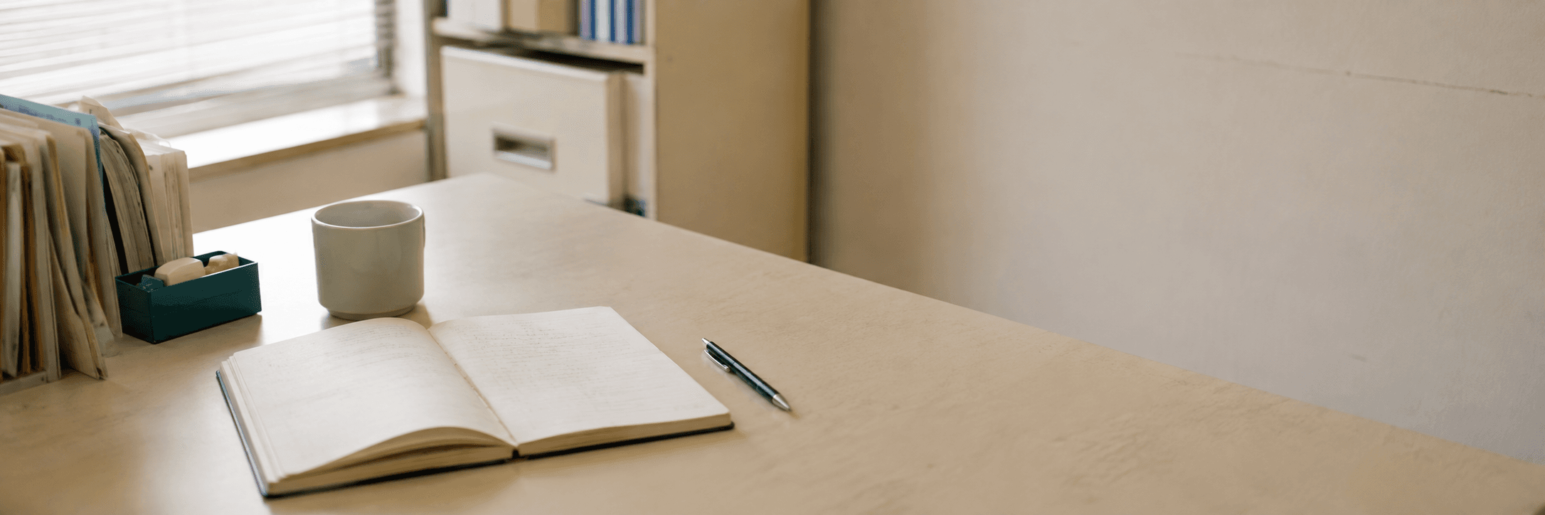 朝の光が入る机の上。書きかけのノートとペン、湯呑み、書類立てが置かれている